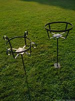 Two black metal pumpkin or plant stands set on grass outdoors. One stand has curled decorative details, the other is simpler. Both have pointed stakes for inserting in ground.