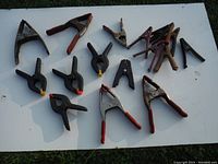 Photo showing various spring clamps on a white surface, including large rusted metal clamps with red handles and smaller plastic clamps with colored tips.