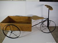 Full side view showing black metal frame, two large spoked rear wheels, one smaller front wheel, wooden seat and handlebar grips, attached large wooden rectangular box with handle on side.