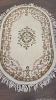 Front view of oval cream wool rug with floral pattern and fringed edges laid flat.