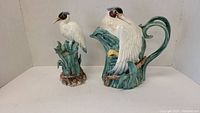 Both Fitz and Floyd ceramic crane bird pieces together showing large pitcher with handle and smaller crane figurine with green leaf and reed bases.