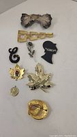 Top view of various brooches including dark metallic bow, gold linked squares, black ampersand, silver flower, black silhouette, gold leaves, and circular pins.