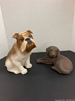 Two ceramic dog figurines displayed on a black surface with a white background; brown and white bulldog sitting and chocolate Labrador lying down.