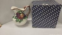 Cream ceramic pitcher with colorful raised winter fruit design and wood-textured handle, with original box beside it