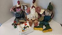 Two large chicken dolls dressed in farmer clothing, green fish doll sitting, stack of white napkins with rooster embroidery, and small black rooster figurine in one shot.