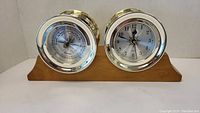 Front view of metal framed clock and barometer mounted on wood base, showing clock face and barometer dial clearly.
