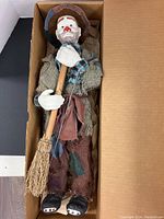Full view of clown doll lying in box showing entire body, clothing, and broom accessory.