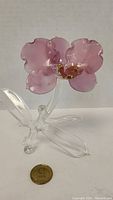 Front view of pink glass orchid figurine showing the pink petals and clear glass stem and leaves with a coin nearby for scale.