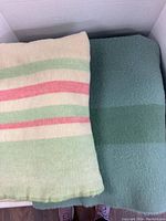 Photo of two folded wool blankets, one off-white with green and red stripes, the other solid green with a darker green stripe, showing texture and condition.
