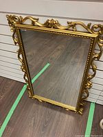 Full view of the rectangular mirror showing the entire gold ornate frame and reflective glass surface on the wooden floor.