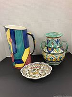 Three items: a geometric pitcher, floral double-handled vase, and a scalloped dish with bird design, against a neutral backdrop.