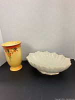Photo showing yellow vase with floral design and white porcelain bowl with scalloped edges and gold trim on black surface.