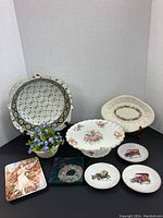 Photo showing an assortment of vintage china including a Grimwades bowl, Coalport cake plate, opalescent flower basket with blue flowers, multiple decorative plates including vintage cars and a maiden artwork, plus a metallic wreath and flower basket vase.