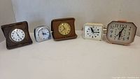 Five vintage travel clocks on flat surface showing front faces and cases.