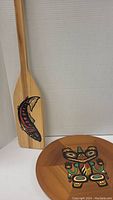 Photo showing the wooden paddle with hand painted salmon design propped behind the round tray with Story of the Bear Mother design visible on top