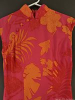 Photo of dress showing vibrant pink and orange floral pattern with mandarin collar, buttons on right shoulder, hanging on white plastic hanger.