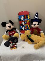 Two 20-inch Mickey Mouse plush dolls, one classic, one Sorcerer Mickey, a 20-inch Pluto plush, fabric pouch, and small decorative box with Mickey images shown on a table.