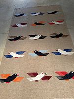 Full view of fringed wool blanket/rug with stylized bird pattern on grey background laid flat on floor.