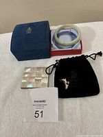 Chinese jade bracelet in red box next to blue velvet Birks watch box, mother of pearl inlaid brass compact, and black Swarovski pouch with dolphin pin.