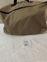 Tan colored closed cargo carrier bag with black handles and zipper, placed on white surface with a MaxSold lot marker '65'