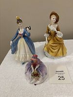 Three Royal Doulton figurines together, two taller standing ladies and one smaller seated figurine, displayed on white cloth
