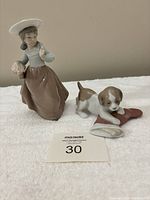 Two Lladro Spain figurines together on a white cloth background: young girl with bonnet and dog with red cloth.