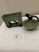 Two Roseville pottery pieces side by side on a white cloth: a rectangular footed planter with open center and a round short ewer with floral raised relief design and handle.