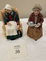 Two Royal Doulton figurines, Forty Winks seated lady and Teatime lady with teapot, with auction lot card in front.