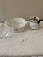 Both pieces of CorningWare Cornflower pattern cookware including square casserole with glass lid and small handles, and teapot with black handle and lid knob shown on white towel