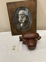Photo of the vintage camera in its brown leather case in front of the old framed photo, showing the carved detail of the wooden frame and oval photo window.