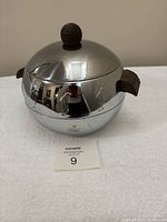 Closed view of a shiny stainless steel spherical insulated server with wooden knobs and handles, showing the seller's reflection.