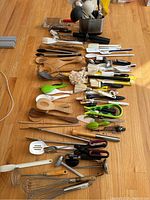 Assorted kitchen utensils spread out on floor including whisks, wooden spoons, spatulas, tongs, peelers, and a metal utensil caddy.