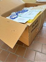 Corrugated cardboard box labeled 'Glass Fragile' containing unassembled chandelier components, packing materials and documents