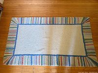 Full view of small rectangular rug lying on hardwood floor showing light beige center and colorful striped border with thin blue inner edge.