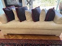 Front view of the cream roll arm sofa showing 7 decorative pillows: 3 matching cream pillows and 4 purple paisley pillows with red fringe.