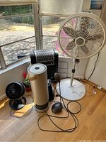 Multiple assorted heating and cooling devices displayed near window including various fans, a portable heater, and an air purifier.