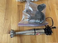 Cuisinart immersion blender showing the stainless steel blending shaft and plastic attachments in a plastic bag on a wooden floor.