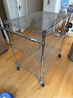 Chrome metal kitchen cart viewed from side angle showing three wire shelves, handle bar, and caster wheels.