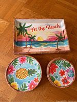 Shows a rectangular tray with a sunset beach scene and 'At the Beach' text, along with two round plates with pineapples and floral designs