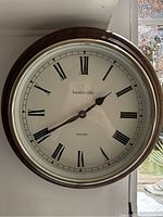 Photo of round wall clock with dark wood frame, white dial with black Roman numerals, black hands, brand Baskerville and model Oxford visible on dial.