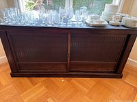 Front view of dark wood console table with black stone top and nail detail on sliding panels. Various glassware and dishes placed on top for staging, not included in sale.