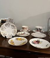 Six plates and teacup sets on table with various floral patterns, gold trimmed, photographed to show full set.