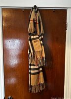 Full view of beige scarf with black, white and red check pattern, fringed ends, hanging against wood door.