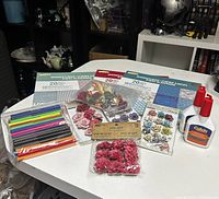 Photo showing assorted floral embellishments in fabric and paper, colorful craft sticks, fiber strands, and two bottles of glue on white table.
