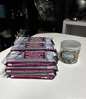 Stack of nine sealed packs of Mortartac Natural Modeling Air Dry Clay with a jar of mixed craft rocks beside them on a white surface.