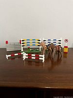 Full view of collection showing four horses and multiple horse jump fences of different color patterns including a flower-decorated green base fence, brick wall obstacle, and yellow no-entry sign post.