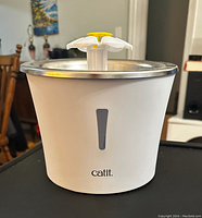 Front view of the Catit pet water fountain showing white plastic base with water level window, stainless steel top with yellow and white flower-shaped water outlet.