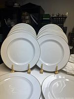 Front view of 12 white Mikasa Doric dinner plates arranged in two rows on plate stands, showing the gold trim and clean surface.