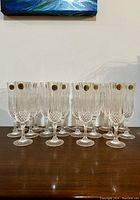 Front view of twenty Cristal d'Arques Longchamp champagne flutes arranged on a wooden surface. Most flutes show original gold brand stickers.