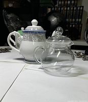 Two teapots on white surface, including a white porcelain teapot with blue decoration and a clear glass teapot with round lid.